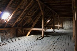Innenansicht eines alten Dachbodens mit freiliegenden Holzbalken und Dachsparren, einem Holzboden und natürlichem Licht, das während des Prozesses des Kellerentrümpelns durch ein Fenster einfällt.